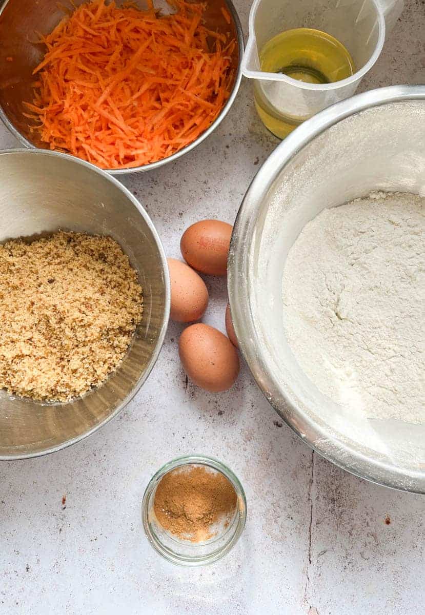 A large silver bowl of flour, a silver bowl of brown sugar, a bowl of grated carrots, three eggs, a jug of oil and a small dish of ground mixed spices. 