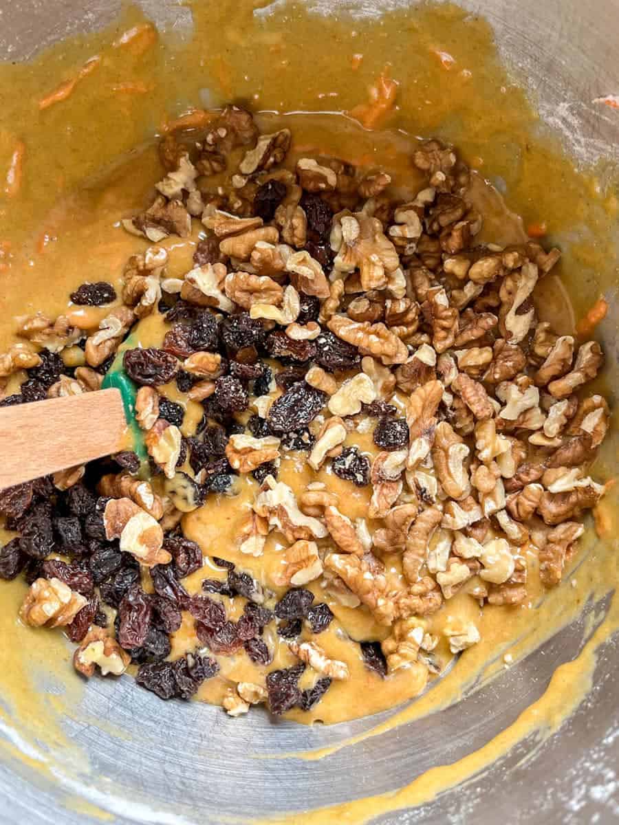 A silver bowl of carrot cake batter with chopped walnuts and raisins.