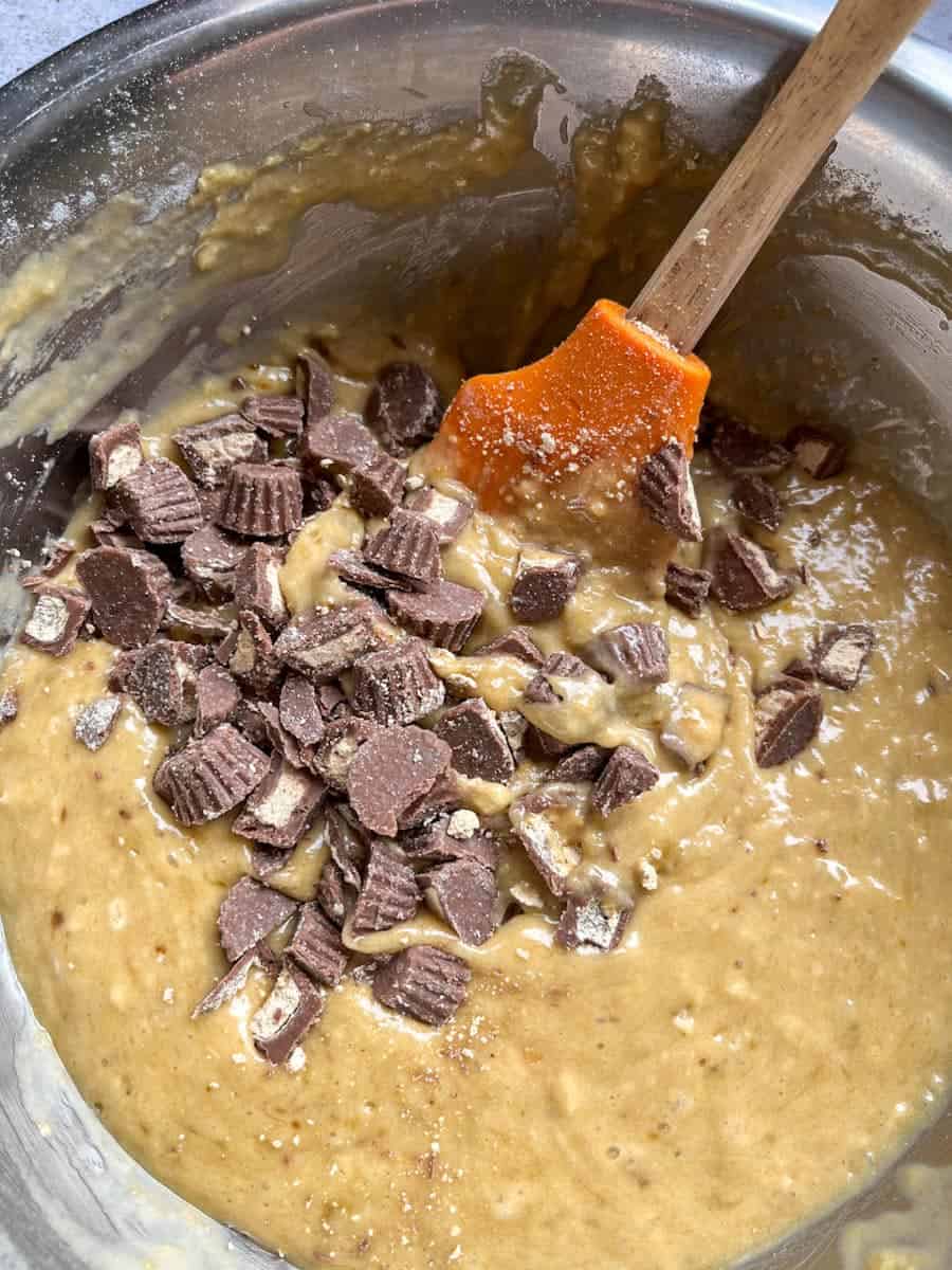 A silver bowl of banana bread batter with chopped peanut butter cups and a wooden spatula.