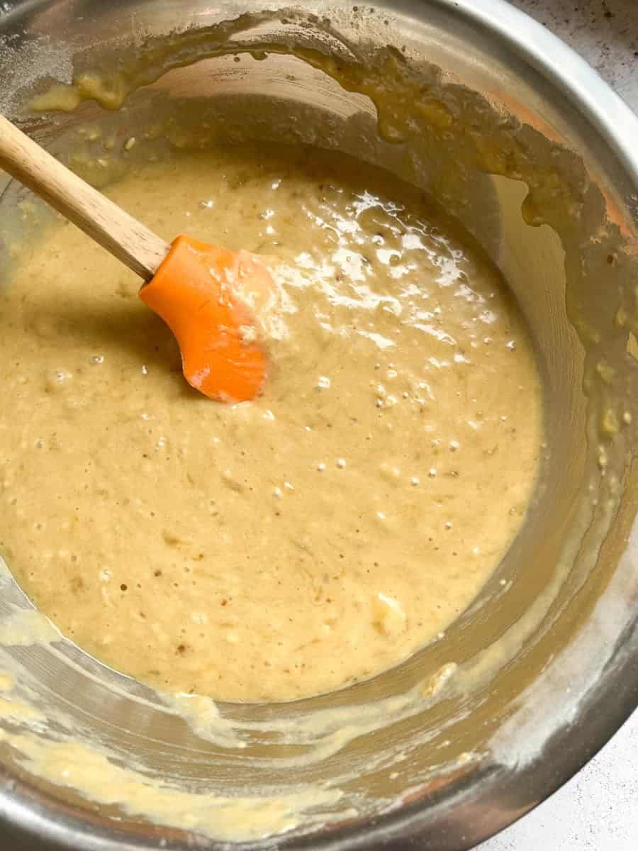 A large silver bowl of banana bread batter with an orange wooden handled spatula.