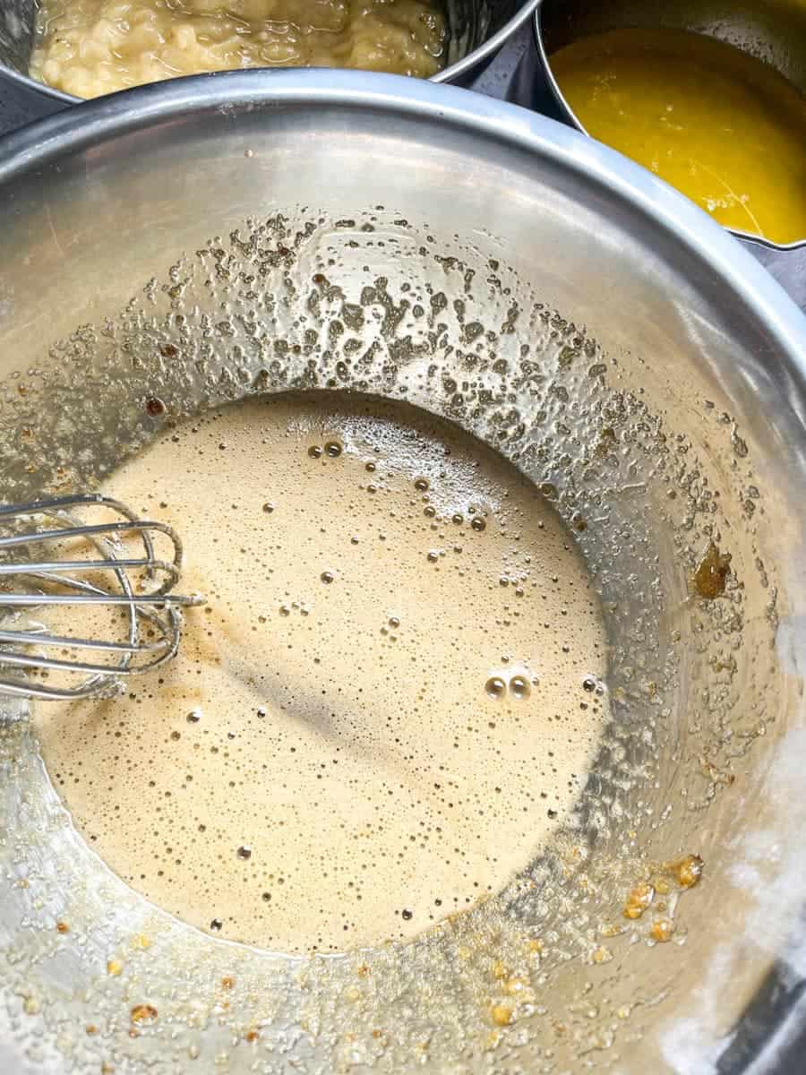 A large silver bowl of beaten eggs, melted butter and sugar with a metal whisk.