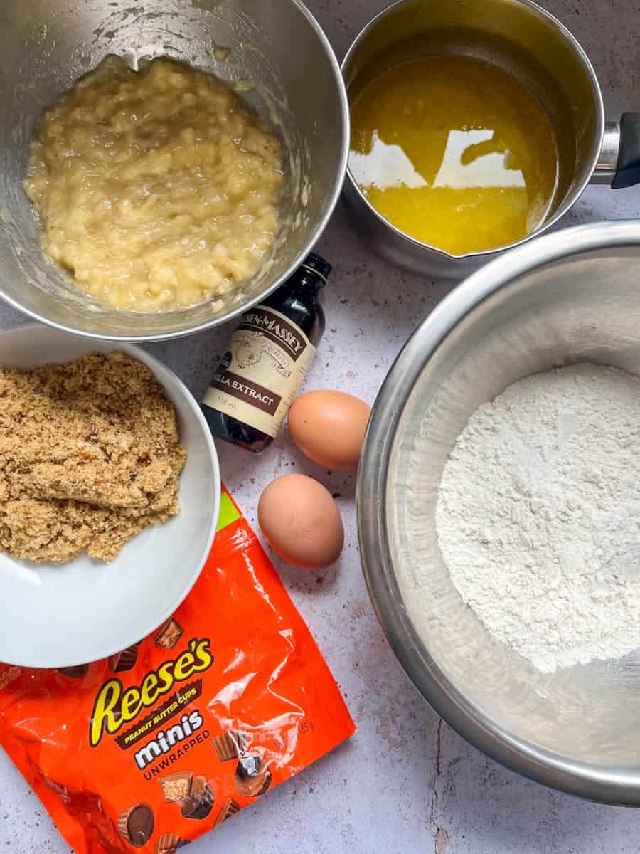 A large silver bowl of flour, a silver bowl of mashed bananas, a white bowl of brown sugar, a saucepan of melted butter, two eggs, a bottle of vanilla extract and a packet of Reeces mini peanut butter cups.