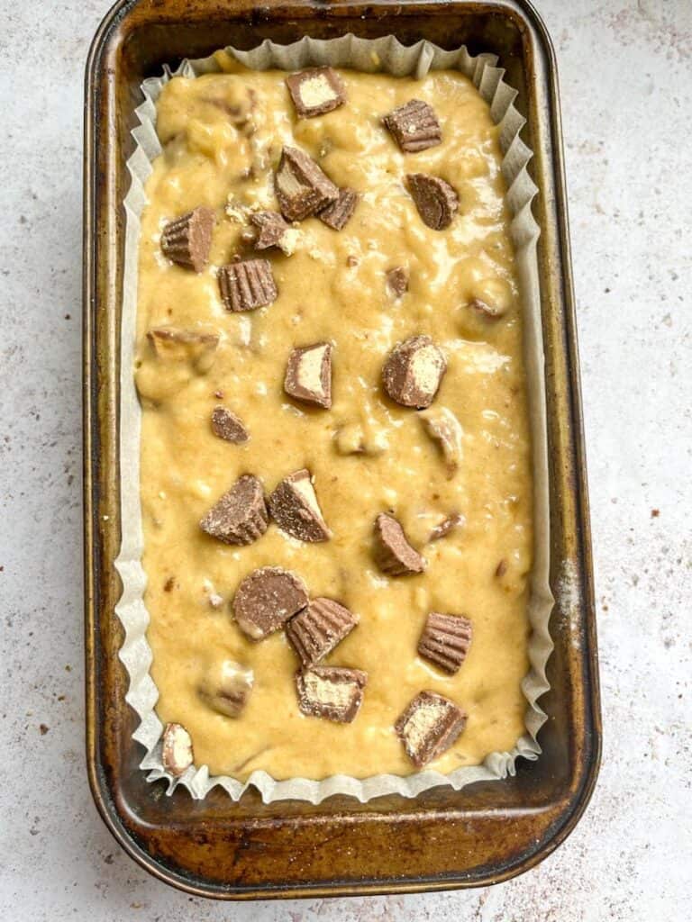 Banana bread batter topped with chopped peanut butter cups inside a loaf tin lined with baking paper.