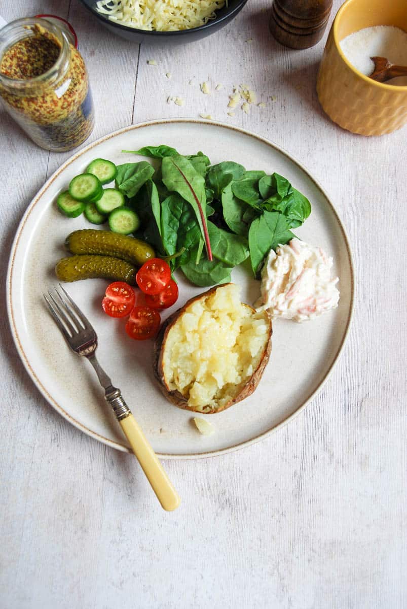 A baked potato with butter on a plate with tomatoes, salad leaves, sliced cucumber, pickled gherkins and coleslaw. A yellow handled silver fork rests on the plate. A jar of mustard, a black bowl of grated cheese and a yellow pot of salt can be partially seen above the plate.