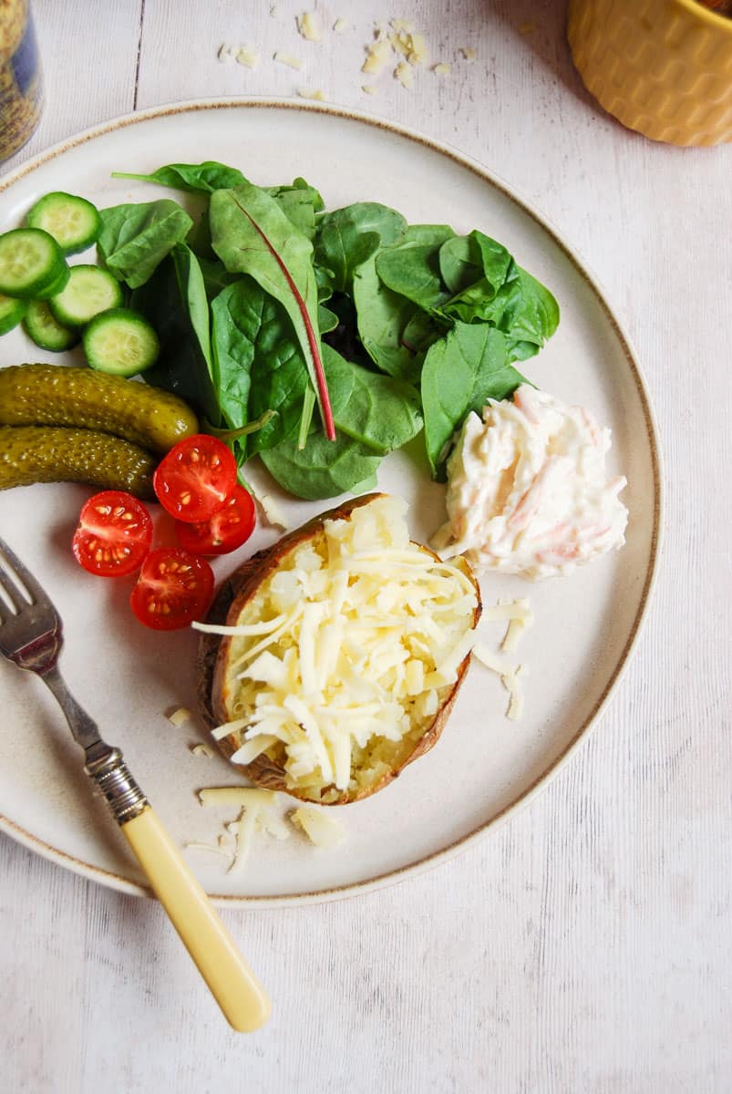 A baked potato topped with grated cheese with salad leaves, sliced cucumber, tomatoes, gherkins and coleslaw. A silver fork with a yellow handle is resting on the plate.