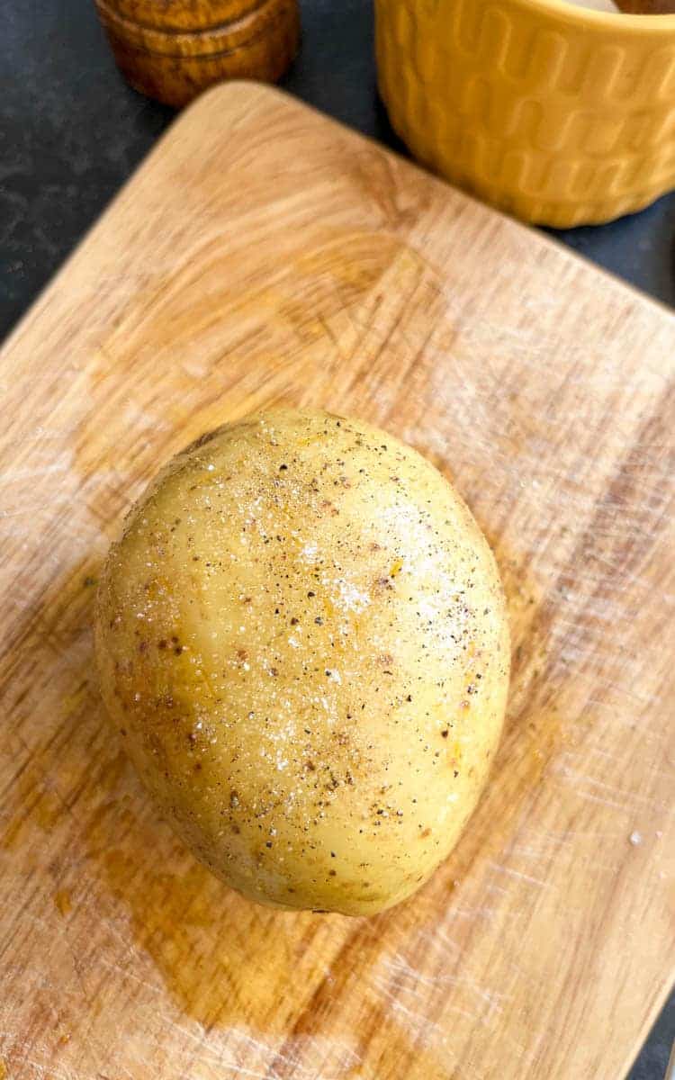 A large baking potato covered in oil and seasoned with salt and pepper on a wooden chopping board.