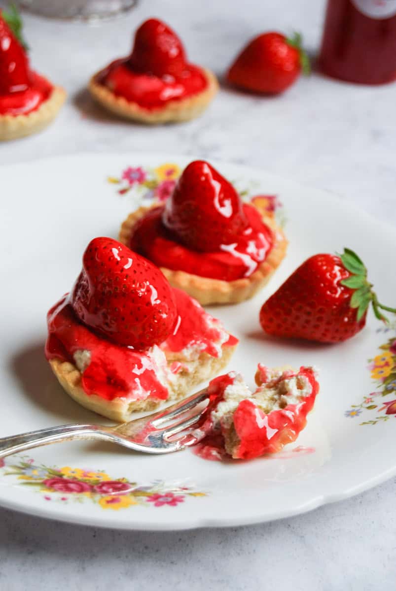 Two strawberry tarts on a floral patterned plate with a fresh strawberry. One tart has been cut with a fork to reveal the cream filling.