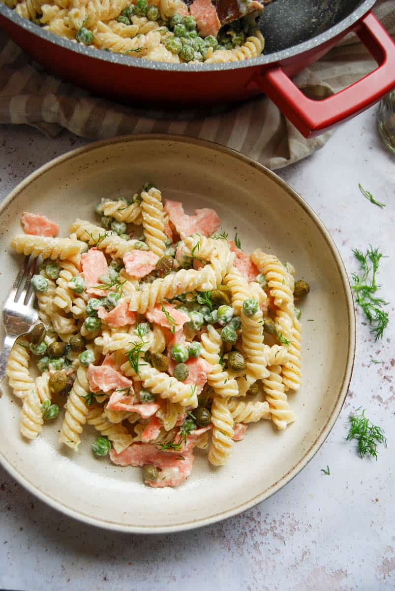 A bowl of Fusilli pasta with smoked salmon, peas and dill in a creamy sauce. A large red pan of pasta sits above the bowl.