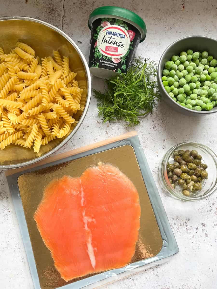 A packet of smoked salmon, a silver bowl of Fusilli pasta, a bowl of peas, a dish of capers, a tub of garlic herb cream cheese and a bunch of fresh dill.