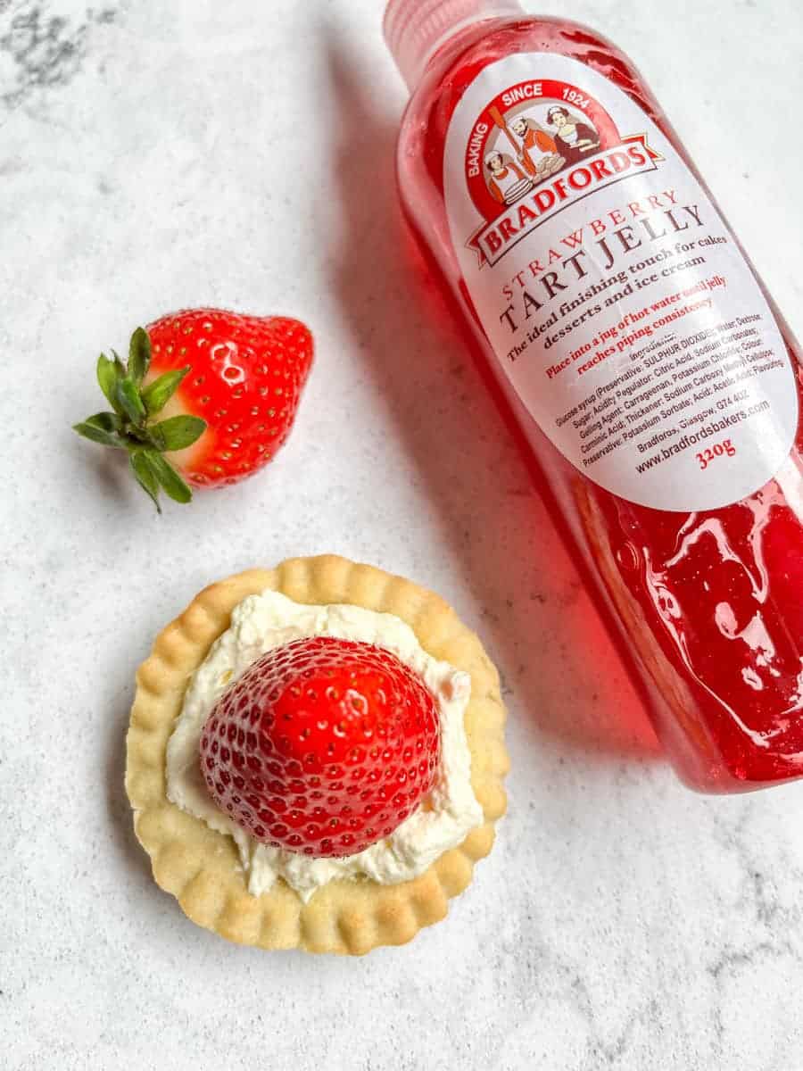 A large strawberry, a bottle of strawberry tart jelly and a small tart shell topped with fresh whipped cream and a fresh strawberry.