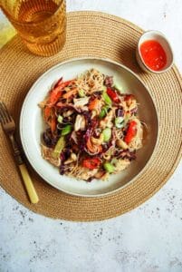 A bowl of chicken noodle salad with sliced vegetables and cashew nuts on a round hessian tablemat, a fork with a yellow handle, a yellow glass of water and a small pot of chilli sauce.