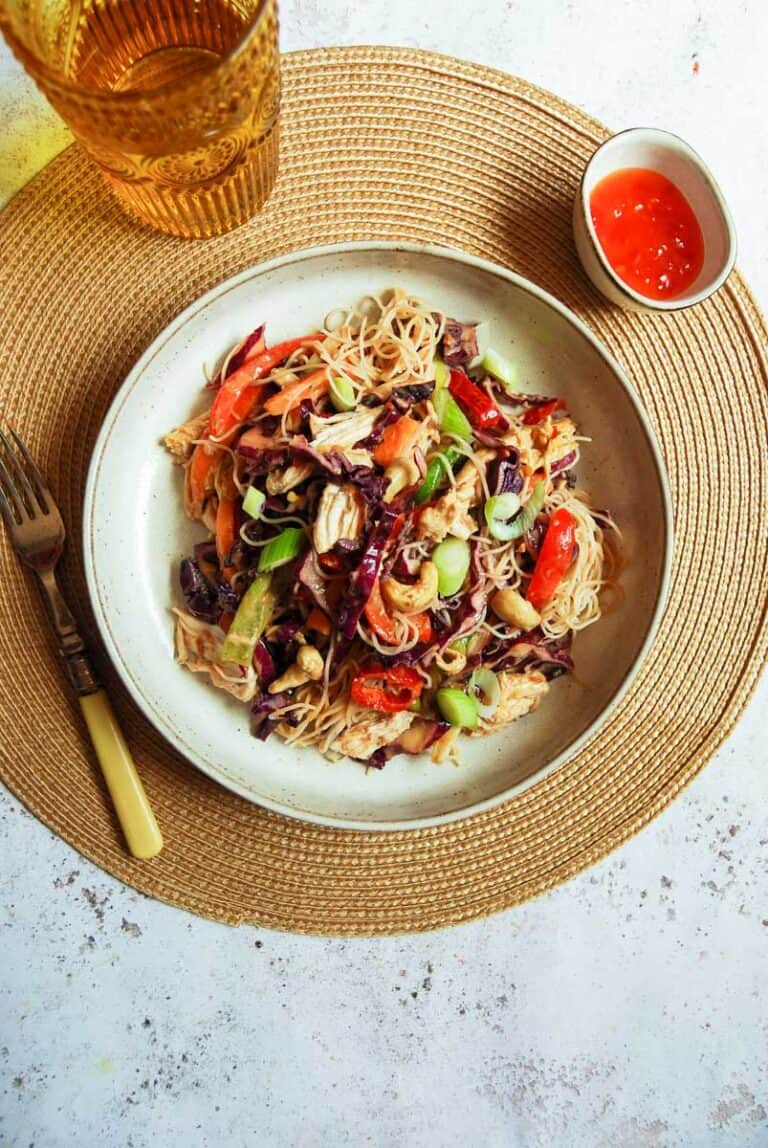 A bowl of chicken noodle salad with sliced vegetables and cashew nuts on a round hessian tablemat, a fork with a yellow handle, a yellow glass of water and a small pot of chilli sauce.
