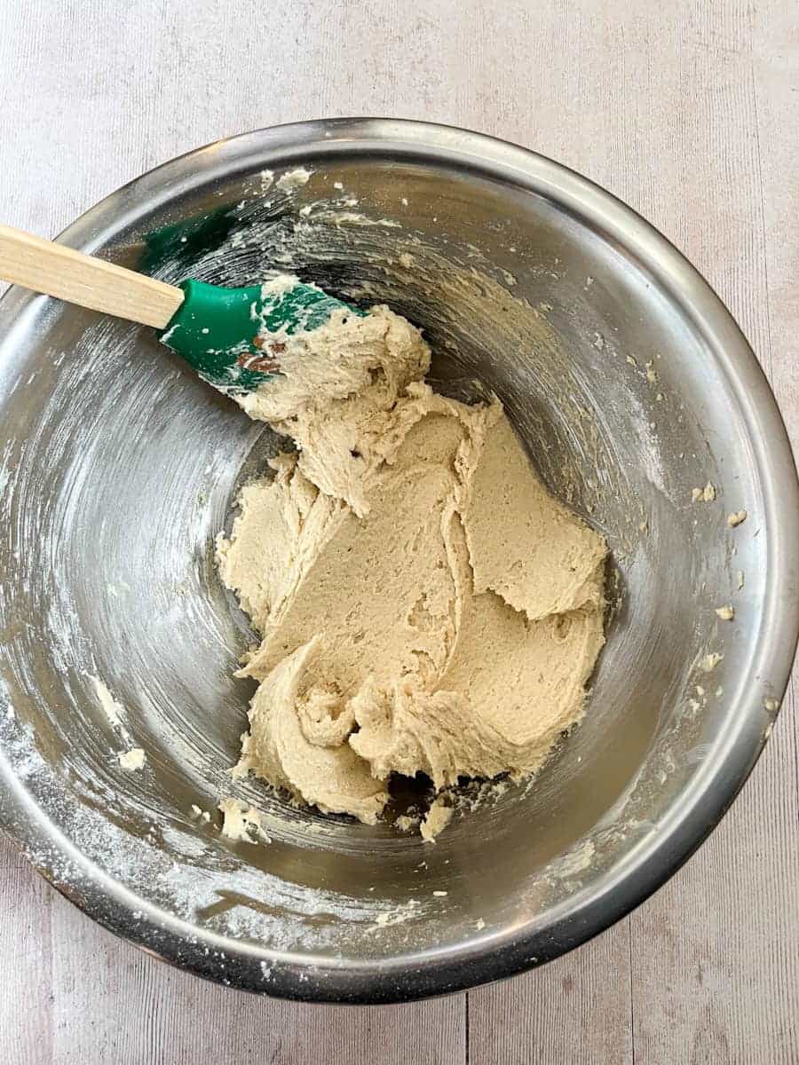 A silver mixing bowl of creamed butter and brown sugar with a green rubber spatula with a wooden handle.