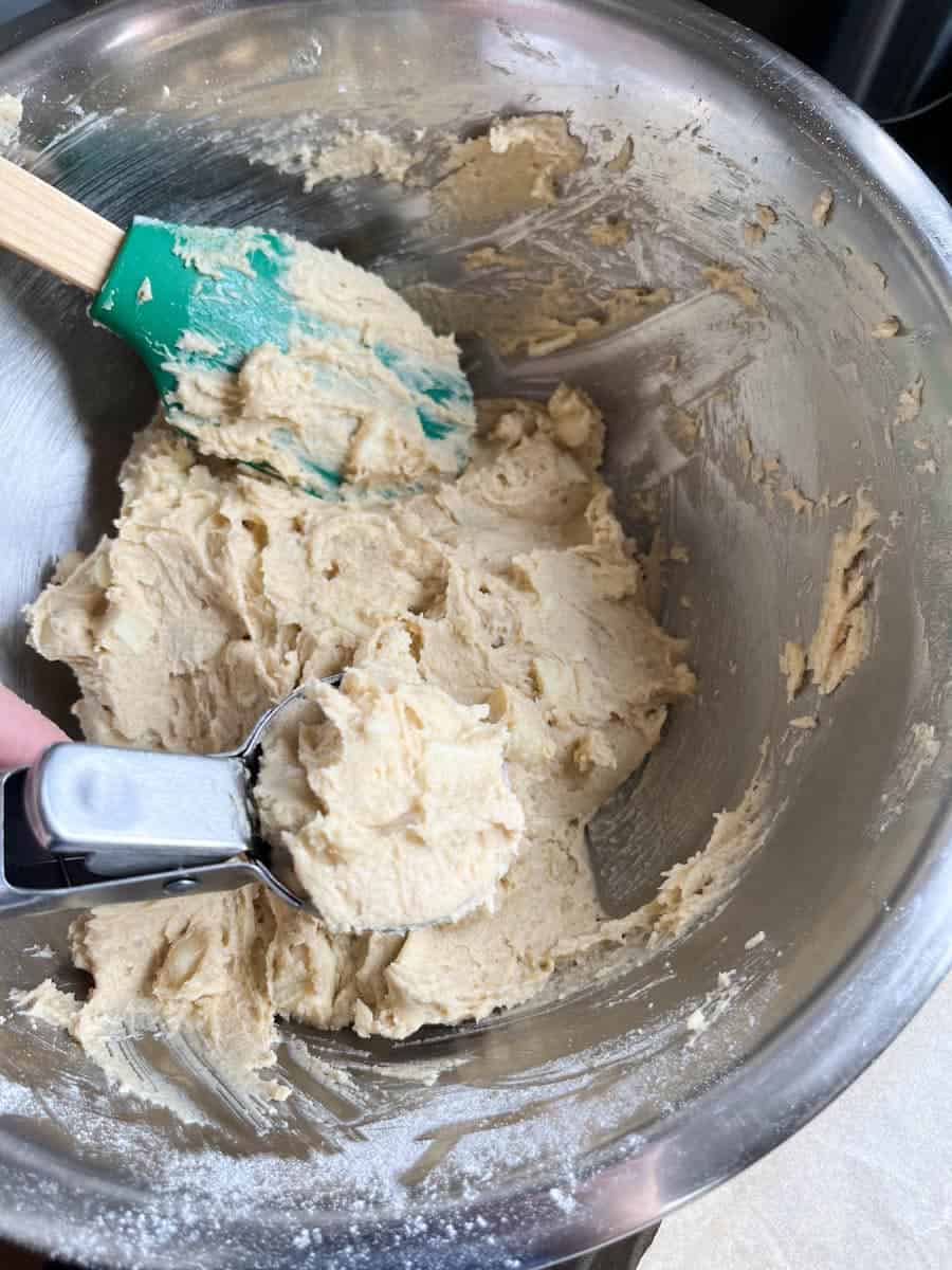A large silver bowl of white chocolate cookie dough with a green rubber spatula with wooden handle. A hand is holding an ice cream scoop with a portion of the cookie dough.