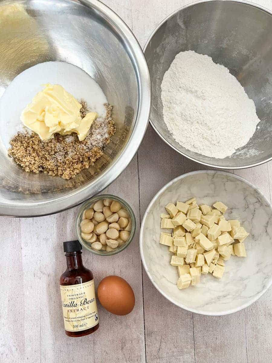 A large silver bowl of butter, white sugar and brown sugar, a silver bowl of flour, a white bowl of white chocolate chunks, a bottle of vanilla extract, an egg and a bowl of macadamia nuts.