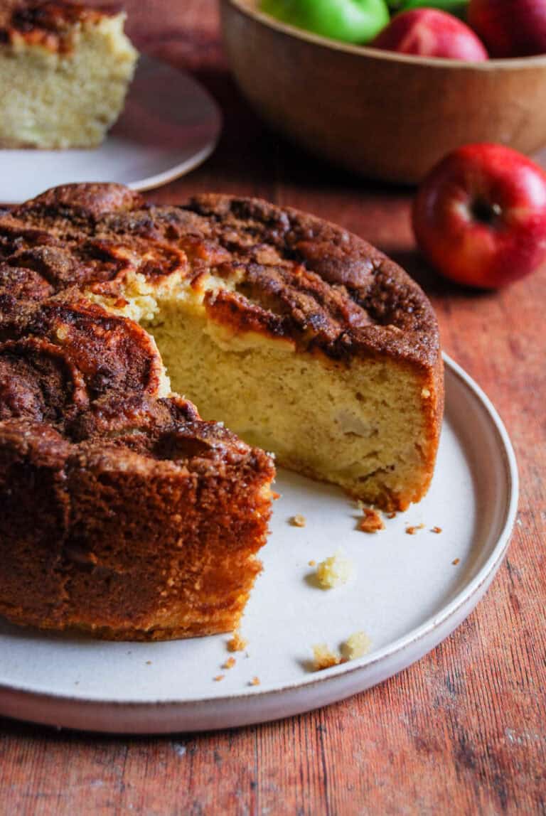 An apple cake with a slice removed to reveal the inside on a beige plate and a wooden bowl of green and red apples.