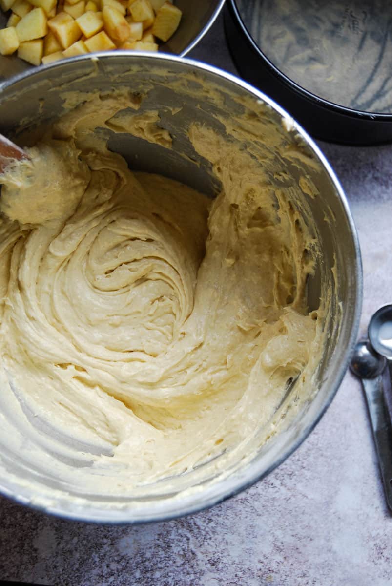 A vanilla cake batter in a silver mixing bowl. A silver bowl of chopped apples, a cake tin lined with baking parchment and a set of silver measuring spoons can be partially seen.
