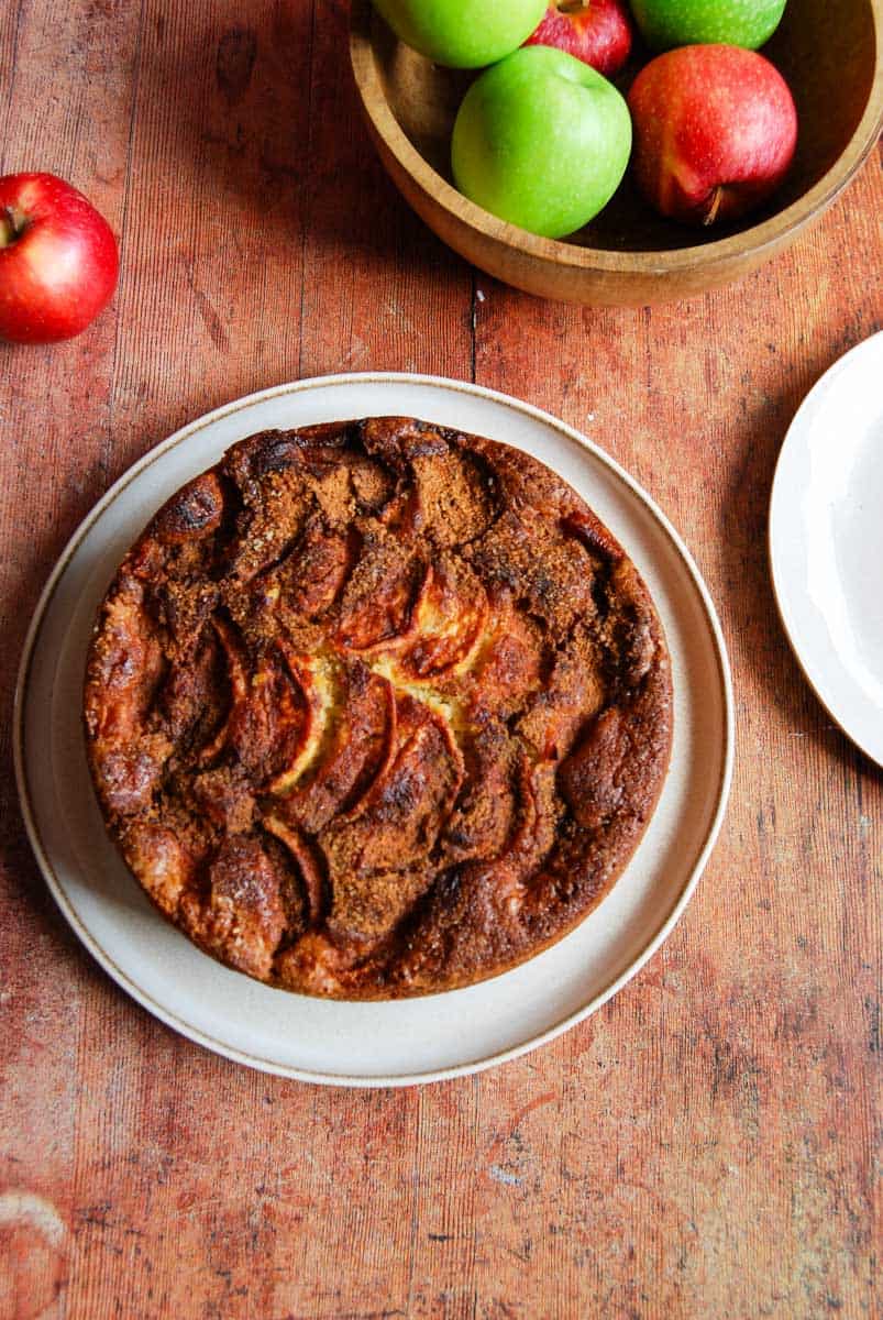 An apple and cinnamon cake on a beige serving plate and a wooden bowl of red and green apples on a wooden tabletop.
