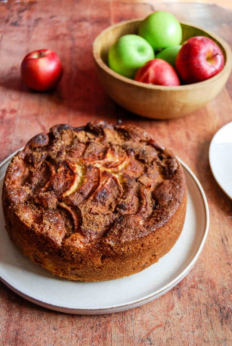 An apple and cinnamon cake on a beige plate and a wooden bowl of green and red apples on a distressed wooden countertop.