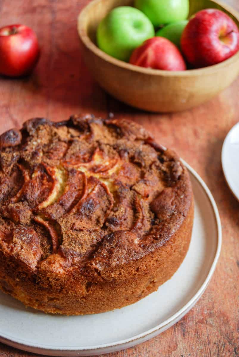 An apple and cinnamon cake on a beige plate with a wooden bowl of red and green apples.