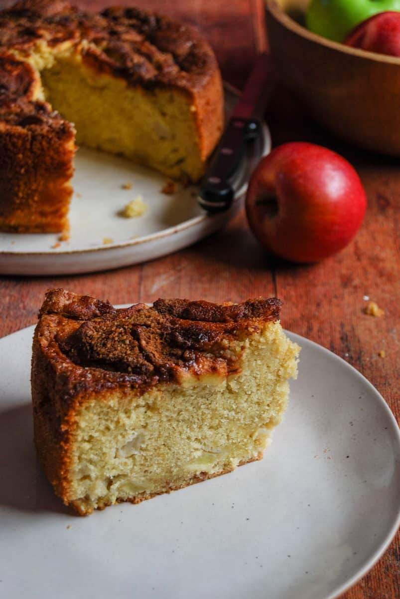A slice of apple cake topped with cinnamon sugar and sliced apples on a white plate. A large apple cake sits on a beige plate in the background with a cutting knife and a bowl of red and green apples.