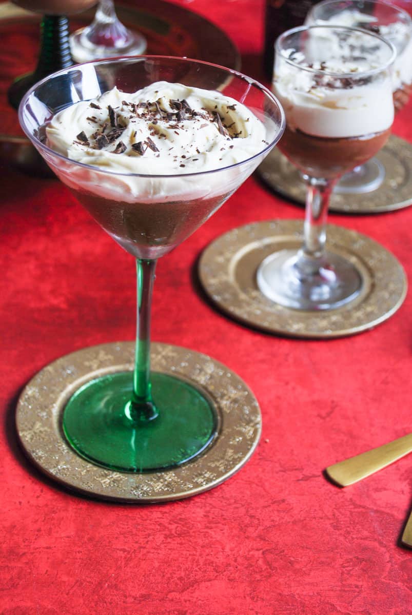 A green martini glass filled with chocolate mousse, softly whipped cream and sprinkled with grated chocolate on a gold coaster. More glasses of chocolate mousse can be partially seen in the background.