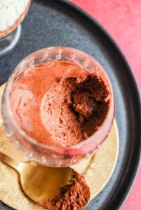A crystal glass of chocolate mousse on a gold coaster and black plate.