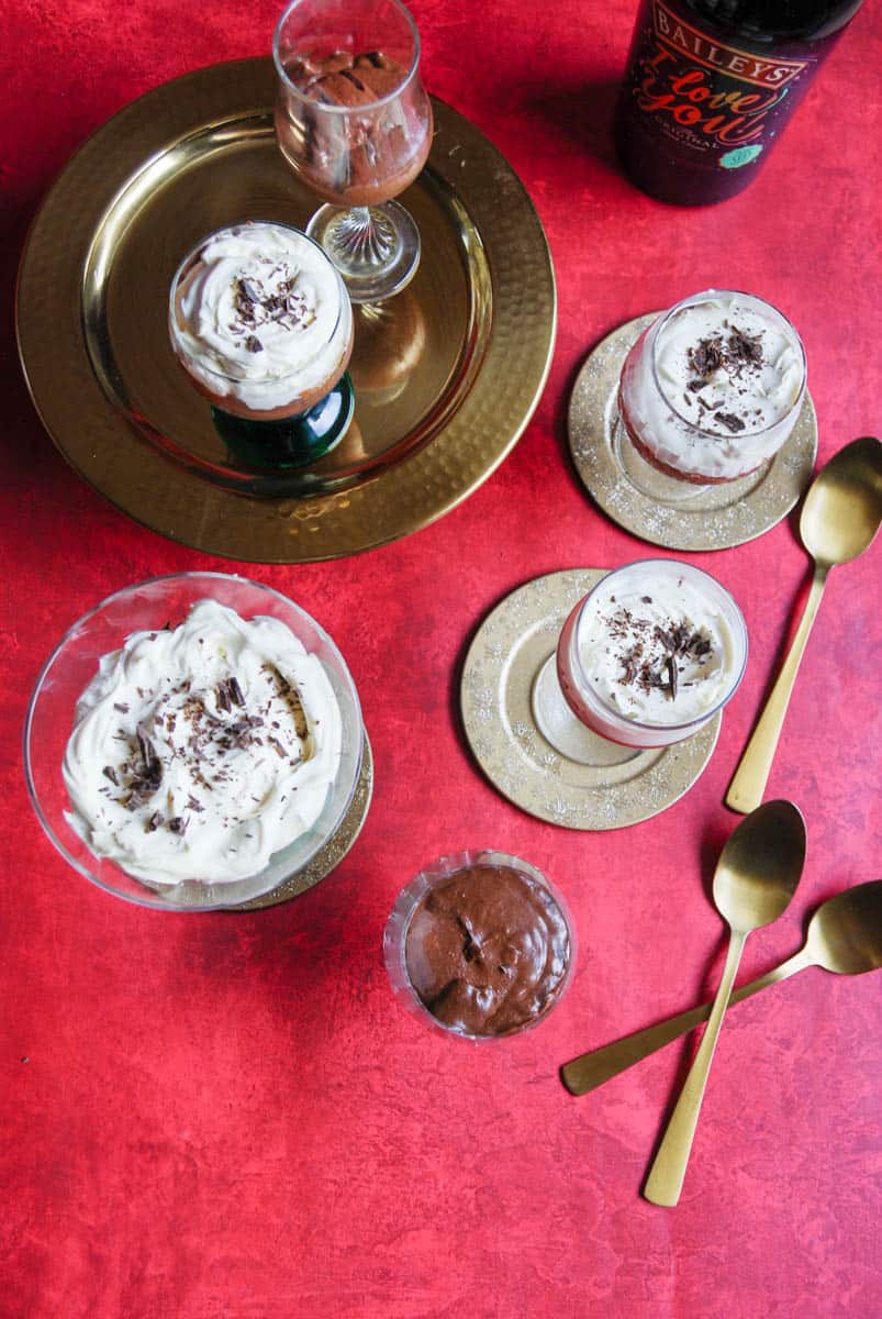 An overhead photo of glasses in various sized filled with chocolate mousse, whipped cream and sprinkled with grated chocolate, three gold spoon and a bottle of Baileys Irish cream liqueur on a red tablecloth.