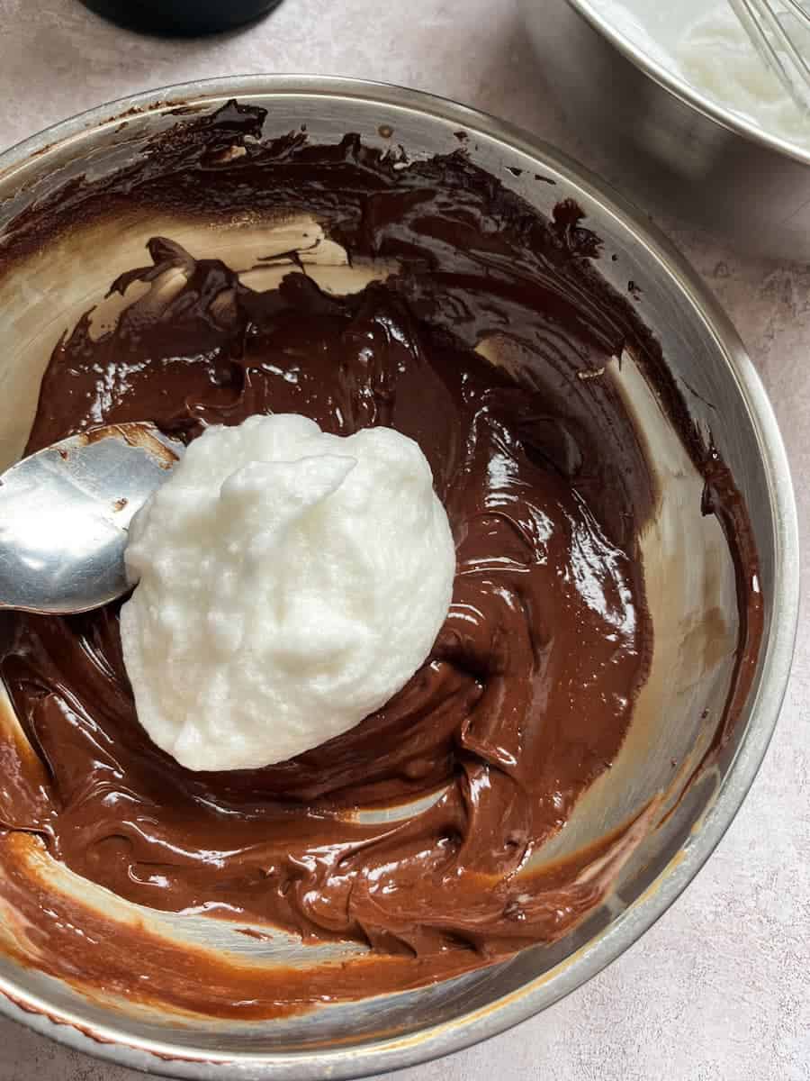 Whisked egg whites being folded into a silver bowl of melted chocolate.