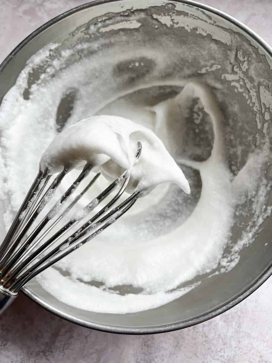 A silver bowl of stiffly whisked egg whites and a silver whisk.