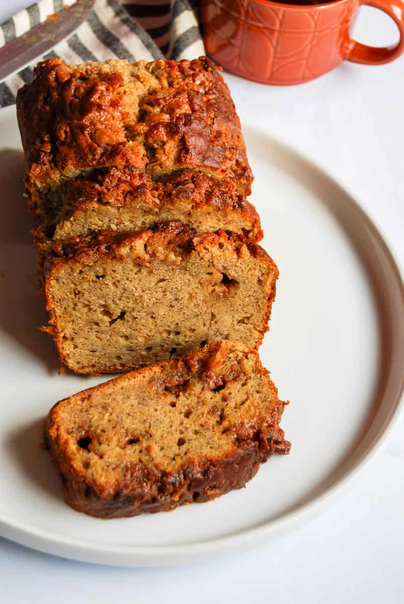 A sliced banana fudge loaf on a white plate with a brown coffee mug and a grey and white striped tea towel.