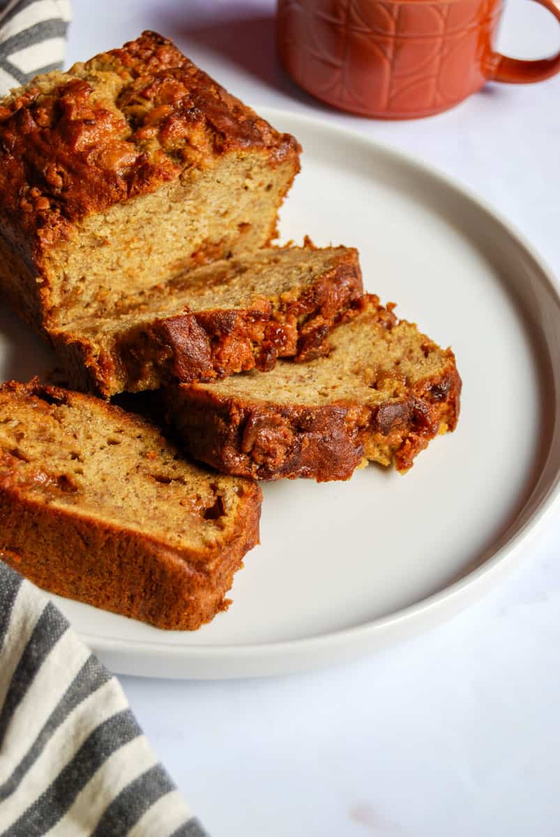 A partially sliced banana loaf on a white plate. A brown mug and a grey and white striped tea towel can be partially seen.
