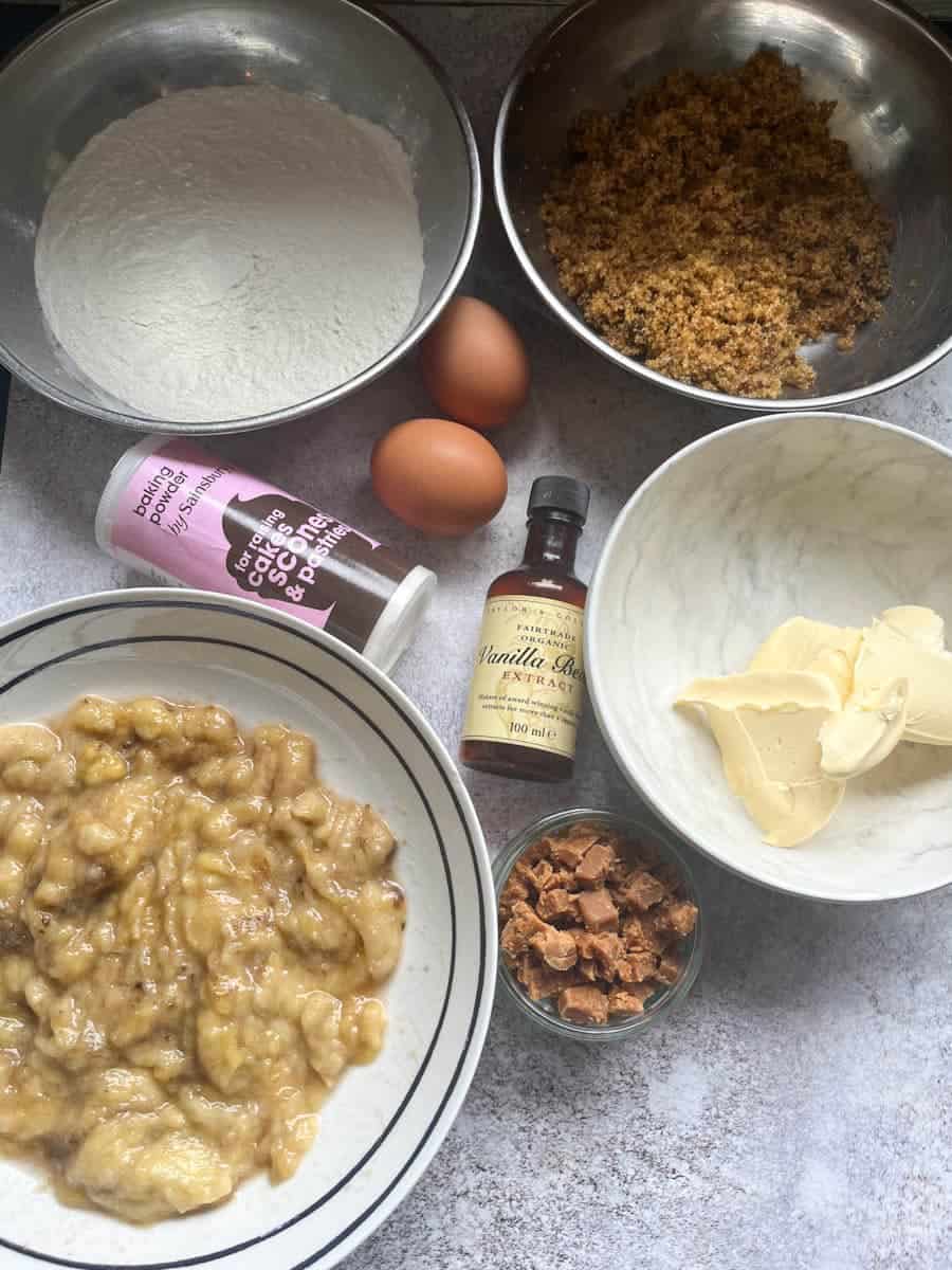 Bowls of flour, brown sugar, butter, mashed bananas, two eggs, a tub of baking powder, a bottle of vanilla extract and a small bowl of chopped fudge pieces.