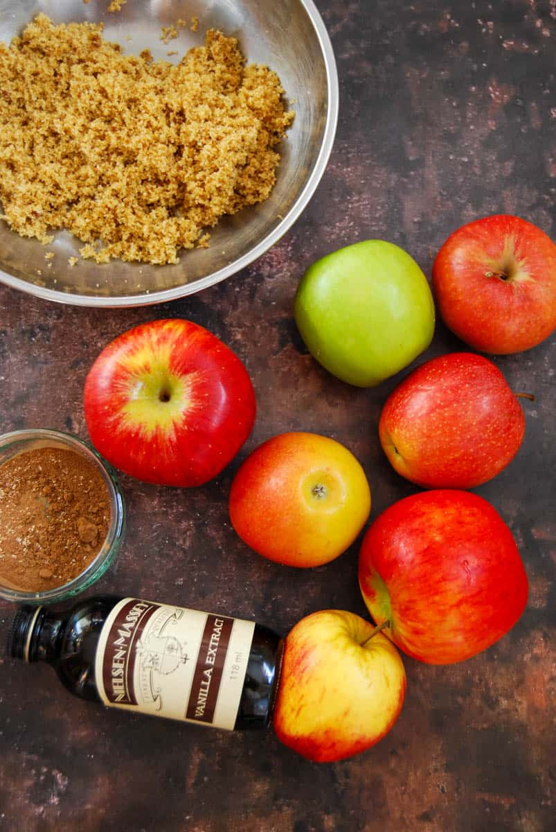 Green and red apples, a bottle of vanilla extract, a silver bowl of brown sugar and a small pot of ground cinnamon and mixed spice on a brown tabletop.