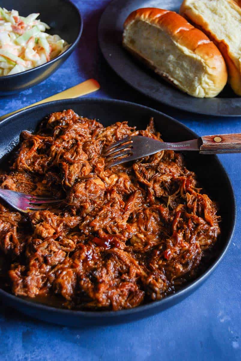 A black bowl of pulled pork with BBQ sauce with two forks, a black plate of hot dog rolls and a bowl of coleslaw.