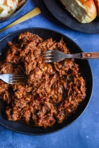 A black bowl of pulled pork coated in BBQ sauce with two forks. A black plate of hot dog rolls and a dish of coleslaw can be partially seen.