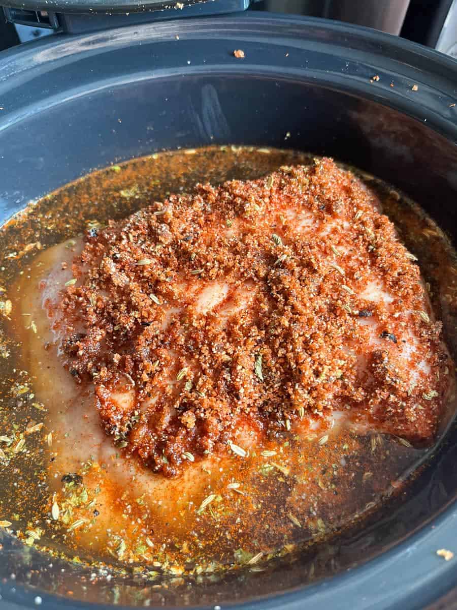 A pork shoulder joint covered in a spice rub and apple juice in a crock pot.