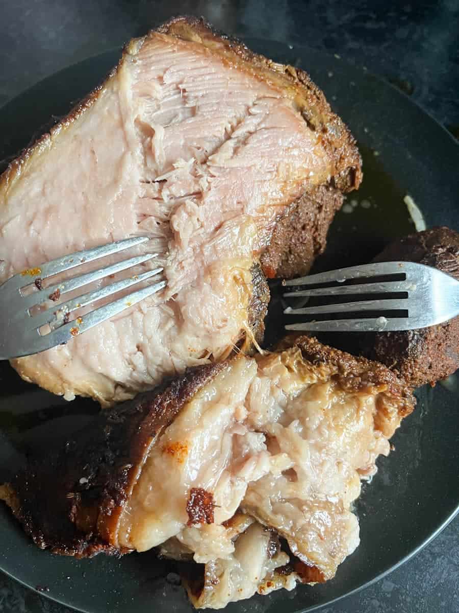 A pork shoulder joint on a black plate with two forks. A layer of fat has been removed from the pork.