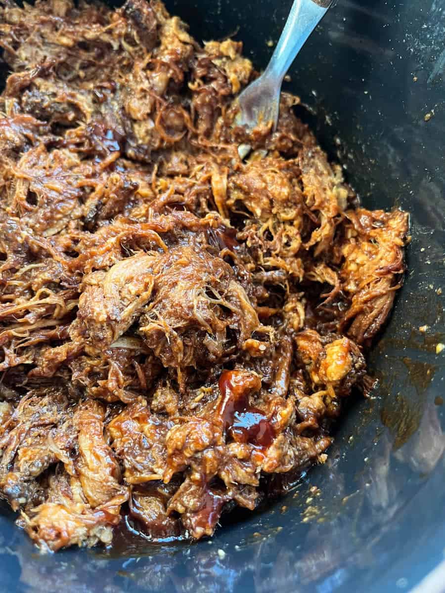 cooked pulled pork in a slow cooker pot with a silver fork.