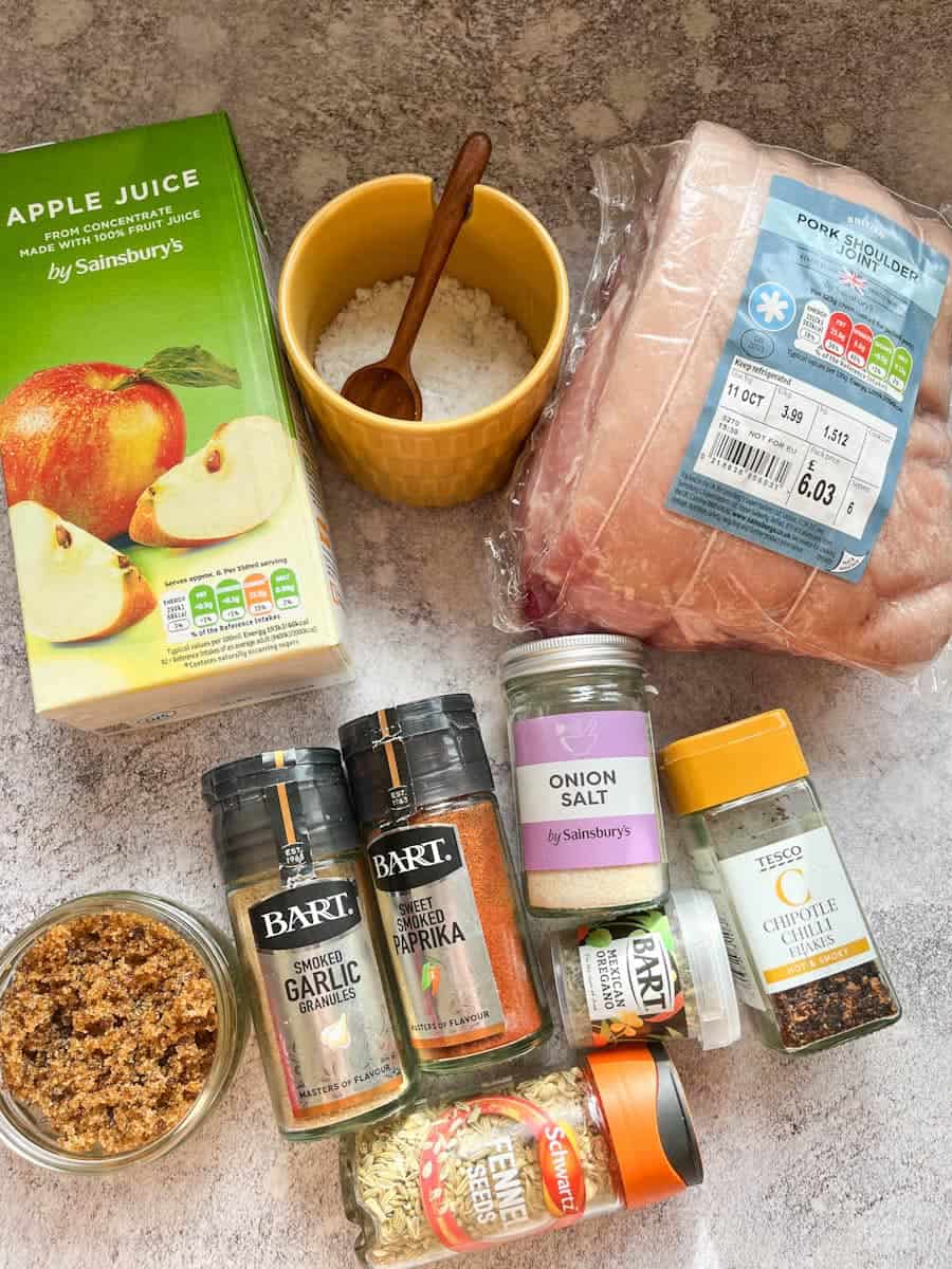 A pork shoulder joint, a carton of apple juice, a yellow pot of salt with a wooden spoon, a pot of brown sugar and various pots of spices.