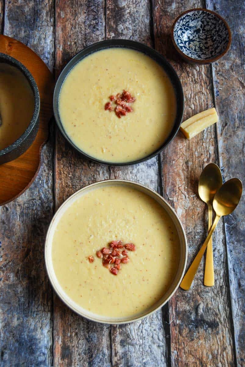 Two bowls of cauliflower soup topped with pancetta, two gold spoons, a blue and brown pot and a parmesan rind.