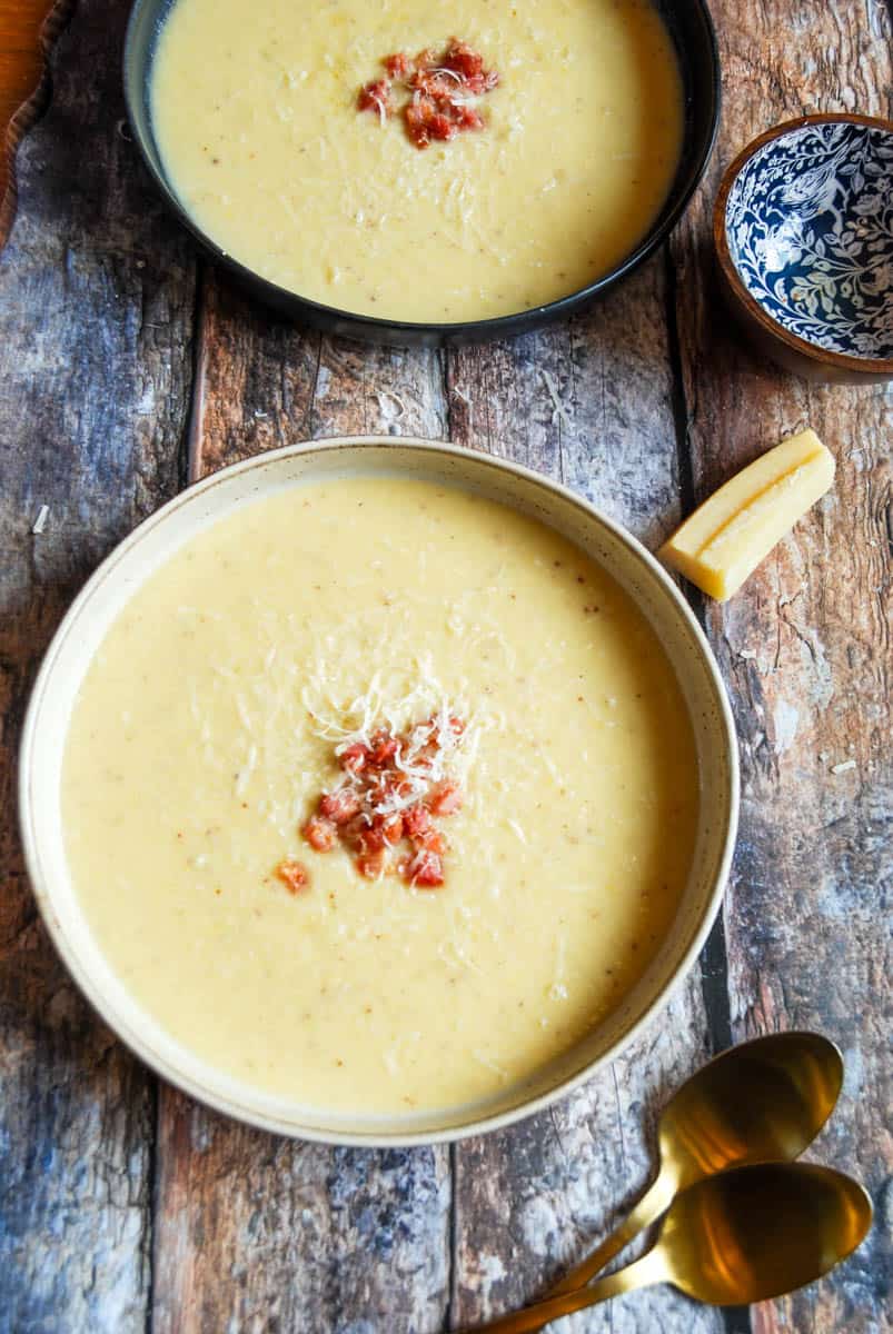 Two bowls of cauliflower cheese soup topped with pancetta and grated parmesan, two gold spoons, a small blue and brown pot and a parmesan rind