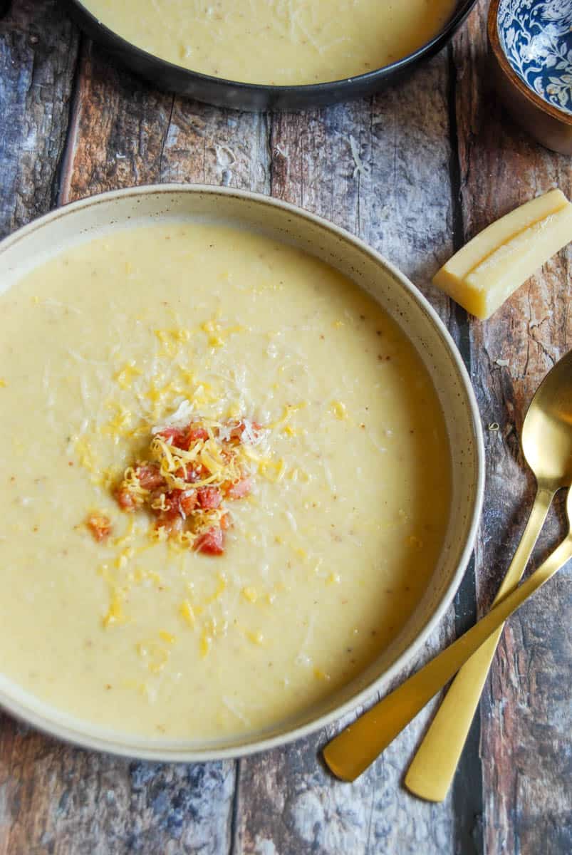 A bowl of cauliflower soup topped with pancetta and grated cheese, two gold spoons and a parmesan rind.