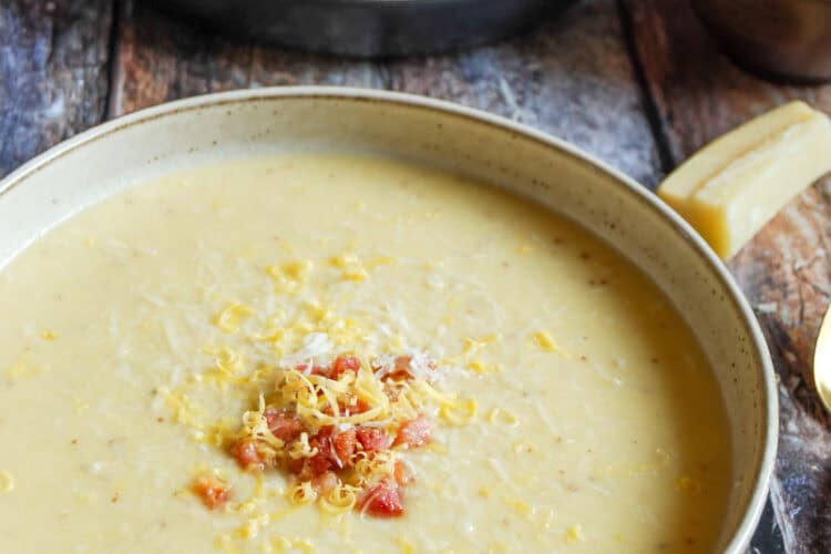 Two bowls of cauliflower cheese soup topped with grated cheese and crispy diced pancetta on a wooden tabletop. Two gold spoons, a blue and brown pot and a parmesan rind can be partially seen.