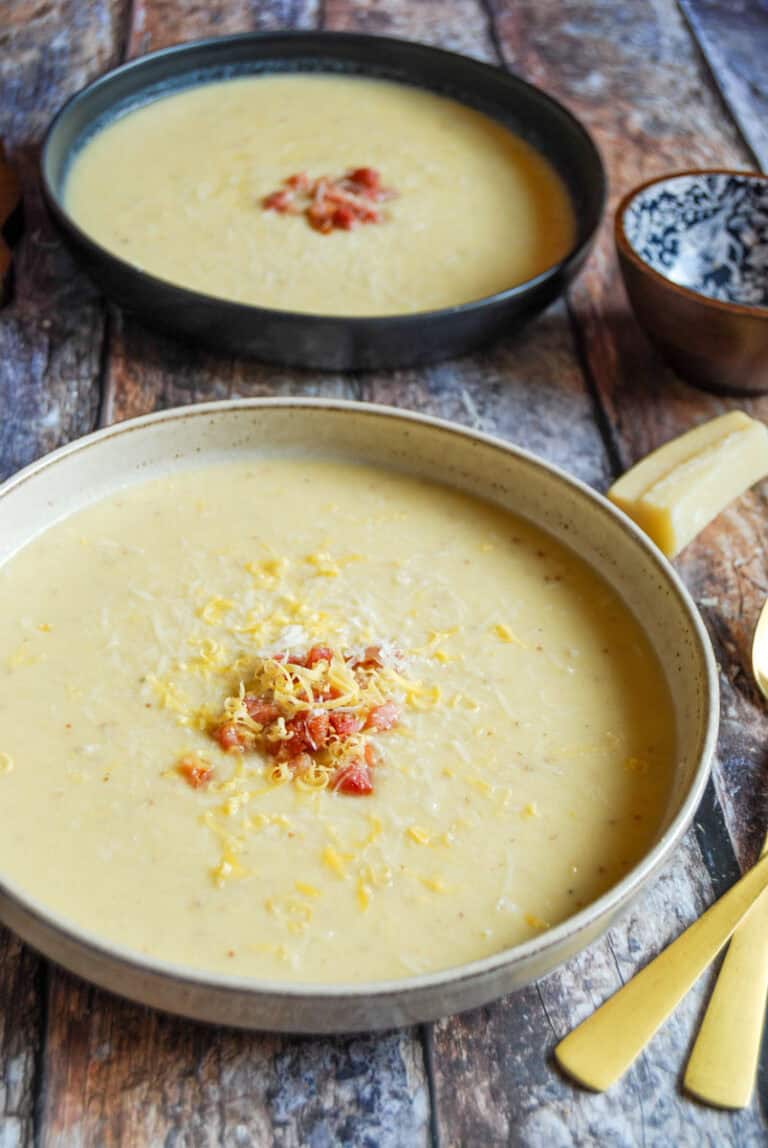 Two bowls of cauliflower cheese soup topped with grated cheese and crispy diced pancetta on a wooden tabletop. Two gold spoons, a blue and brown pot and a parmesan rind can be partially seen.