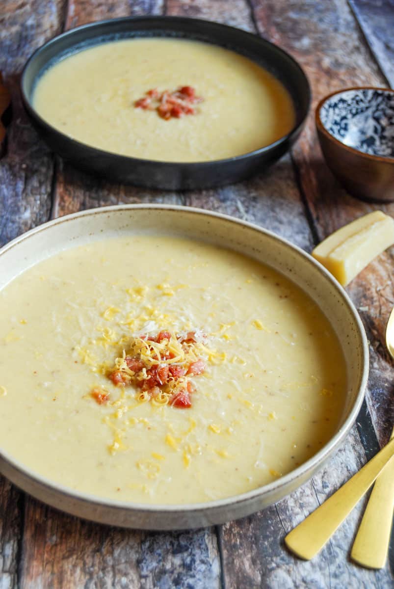 Two bowls of cauliflower cheese soup topped with grated cheese and crispy diced pancetta on a wooden tabletop. Two gold spoons, a blue and brown pot and a parmesan rind can be partially seen.