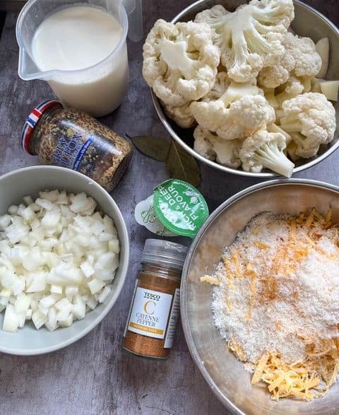 Bowls of chopped onion, cauliflower florets, grated cheese, a jug of milk, a jar of wholegrain mustard, two stock pots and a jar of cayenne pepper.