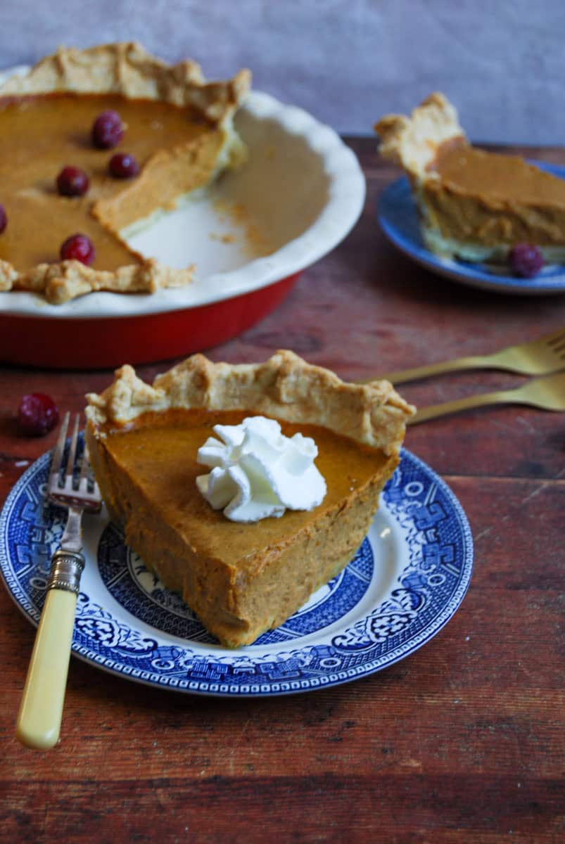 A slice of pumpkin pie topped with whipped cream on a blue willow pattern plate with a silver fork. A large pumpkin pie topped with sugared cranberries with a second slice of pie is in the background.