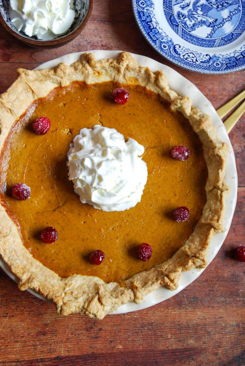 A pumpkin pie topped with whipped cream and decorated with sugared cranberries, a wooden bowl of whipped cream and a blue willow pattern plate.