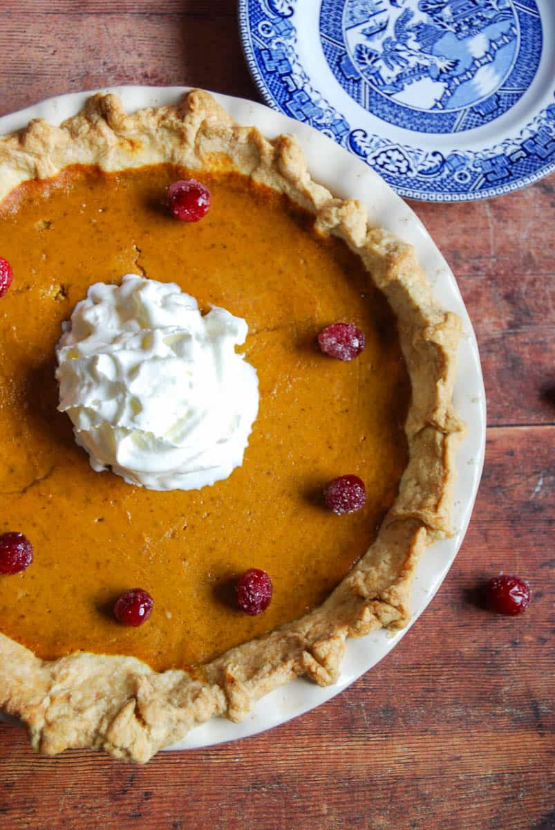 A pumpkin pie topped with whipped cream and sugared cranberries and a blue willow pattern plate.